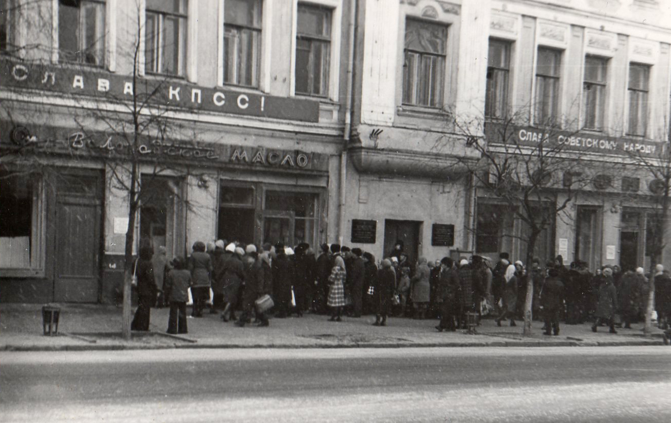 Очередь в фирменный магазин «Вологодское масло», комично сочетающаяся с лозунгами, вывешенными на здании к празднику 7 ноября, 1970-е годы